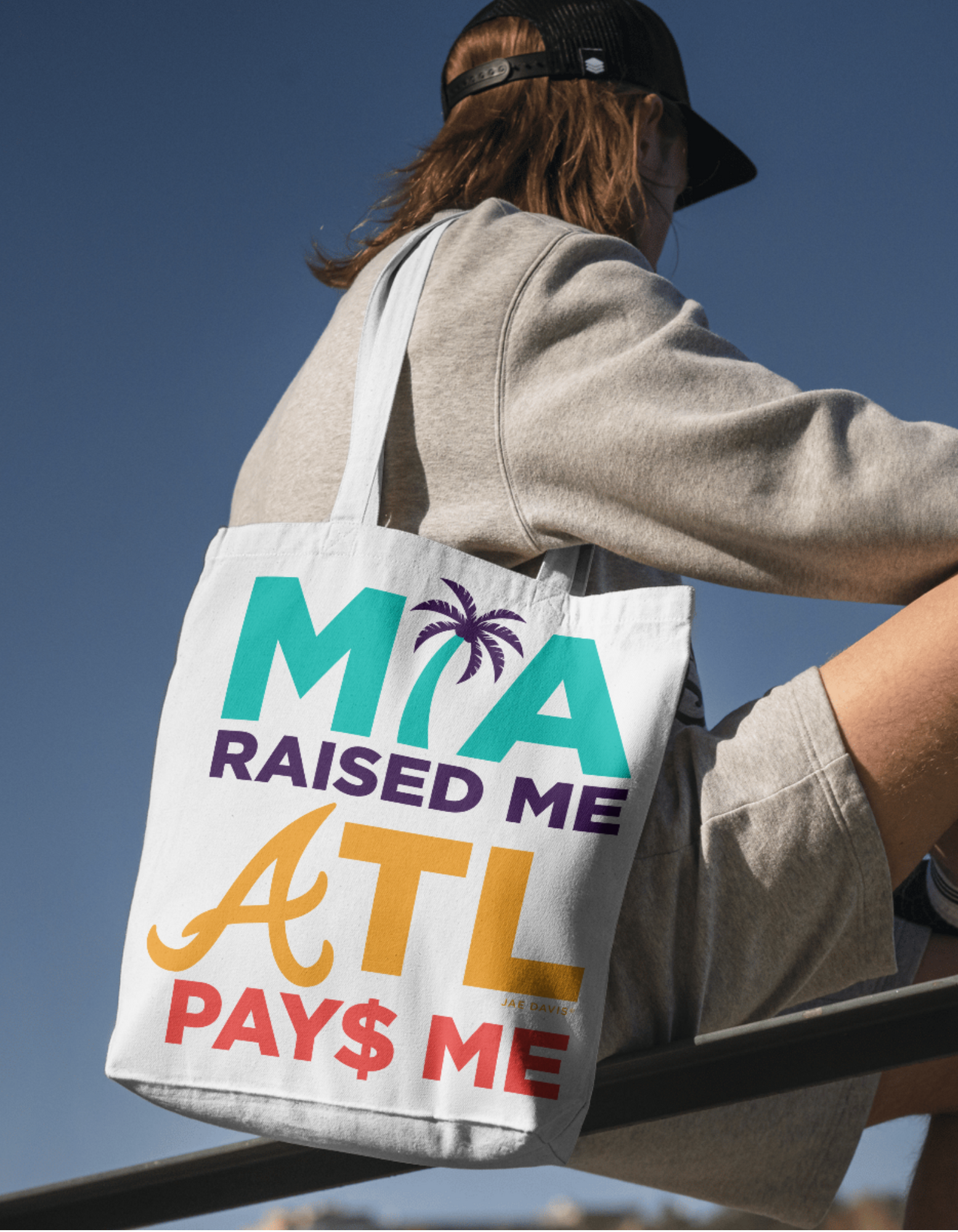 Person holding a tote bag with 'MIA Raised ME ATL PAY$ ME' text against a clear blue sky.