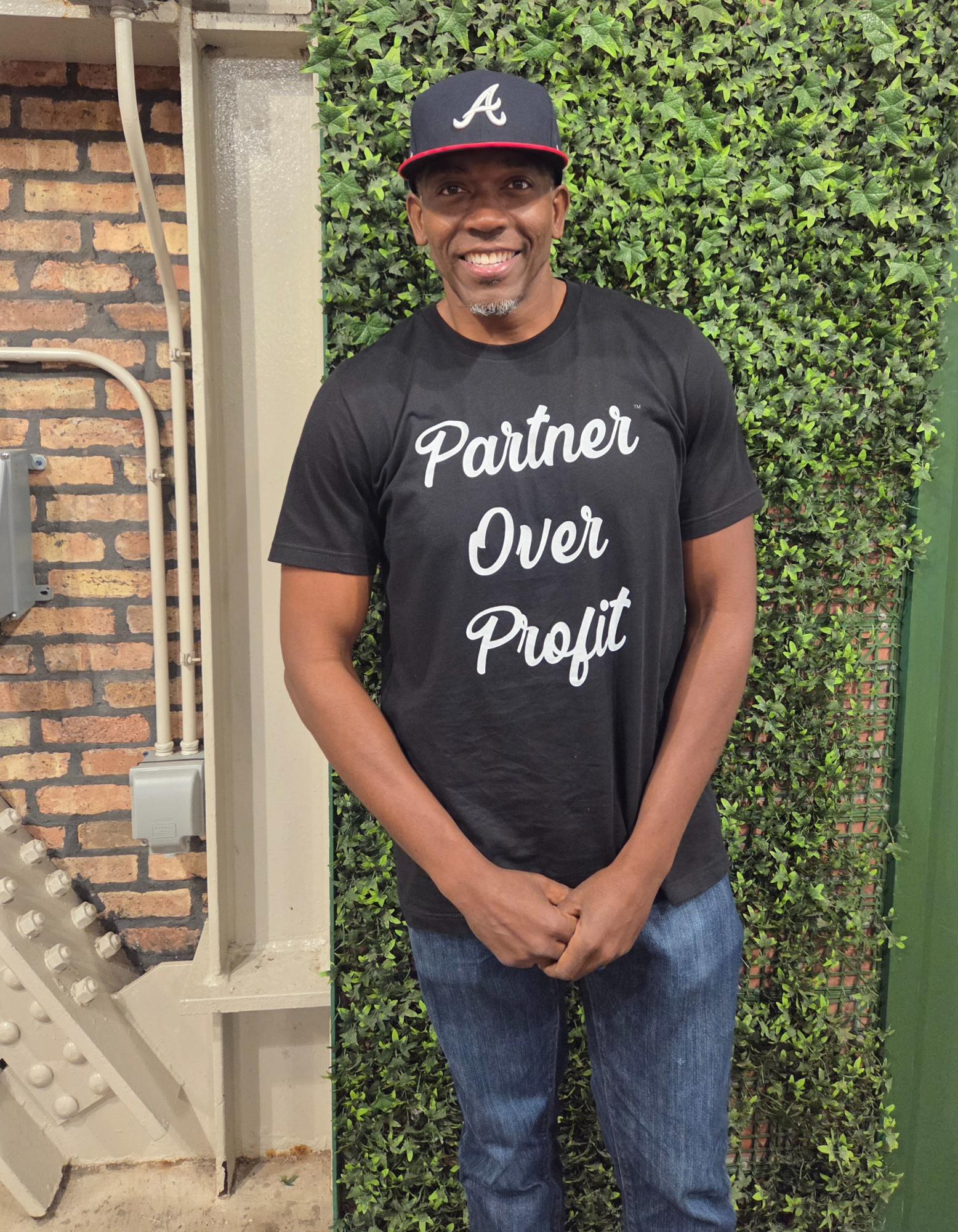 Person wearing a black t-shirt with 'Partner Over Profit' text and a cap, standing against a green wall.