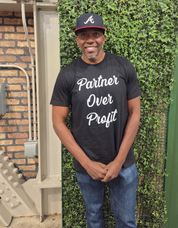 Person wearing a black t-shirt with 'Partner Over Profit' text and a cap, standing against a green wall.