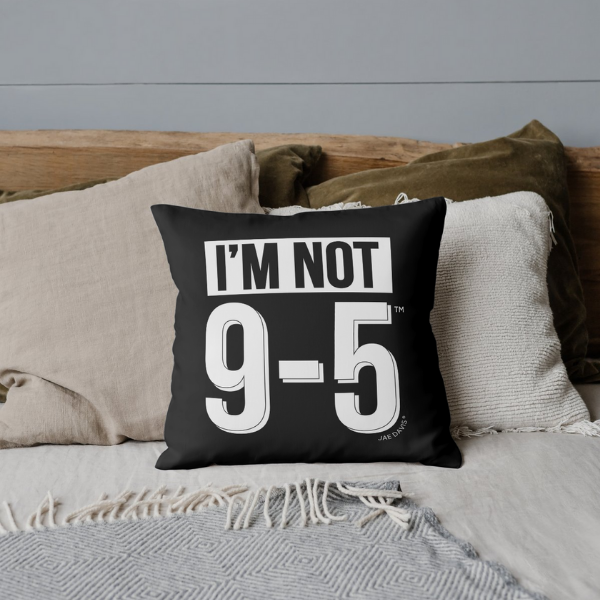 Black pillow with 'I'm Not 9-5' text on a bed with neutral-colored pillows and a gray wall.