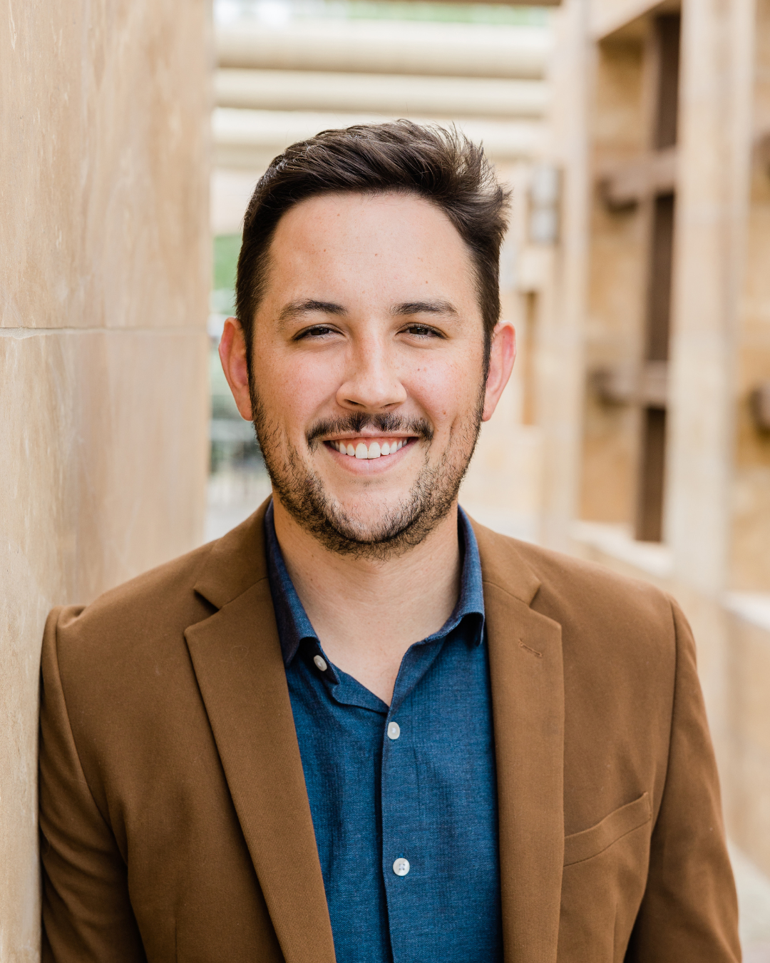 Man wearing a brown blazer over a blue shirt, standing against a beige wall.