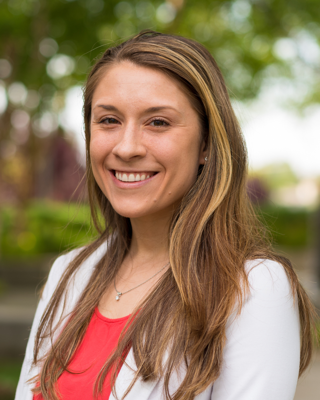 Woman with long brown hair smiling outdoors with a blurred natural background